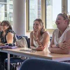 Lachende Teilnehmerinnen bei einem berufsbegleitenden Studium. Sie sitzen an Tischen mit Tablets und Notizbüchern.