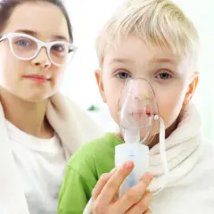 Dziecko z maską inhalacyjną, wspierane przez siostrę w okularach. Dzieci przechodzą inhalacje z soli fizjologicznej.