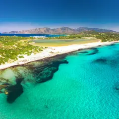 Widok z lotu ptaka na turkusowe morze i piaszczyste plaże w okolicach Cagliari. W oddali widać góry i zatokę z łodziami.