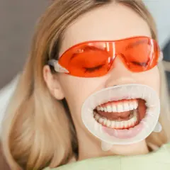 Uśmiechnięta blondynka w pomarańczowych goglach i rozwieraku dentystycznym. Sprawdź, jaka jest higienizacja zębów cena i zadbaj o swój uśmiech!