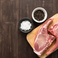 Deux belles côtes de porc crues sur une planche en bois, prêtes pour une cuisson au four, avec du sel et du poivre en grains à côté.