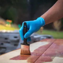 Main tenant une brosse, applique une lasure sur des planches de bois à l'extérieur.