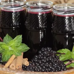 Sok z czarnego bzu – przepis babci. Trzy butelki ciemnego soku, świeże owoce czarnego bzu, listki mięty i laski cynamonu na drewnianej tacy.