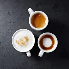 Trzy białe filiżanki z kawą na ciemnym tle. Jedna to espresso, druga to kawa macchiato z pianką, a trzecia to latte.