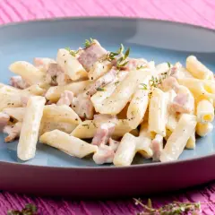 Penne pasta with creamy sauce and diced ham, garnished with thyme, on a blue plate.