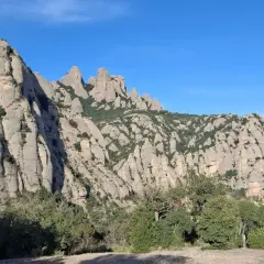 Imponentes rocas de Montserrat bajo un cielo azul, con vegetación verde y la creu de Sant Miquel en la distancia.
