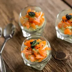 Trois verrines de saumon, avocat et caviar, garnies d'aneth, sur une table en bois. Deux cuillères argentées sont posées à gauche.