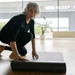 Une femme prépare son tapis pour faire du sport chez soi. Elle est à genoux, déroulant le tapis gris sur le sol en bois.