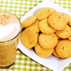 A glass of coffee with a heart-shaped foam art next to a plate of golden brown, round cookies.