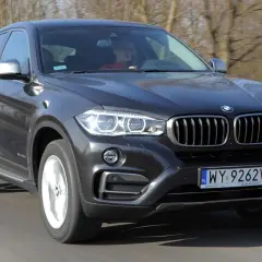 Ciemnoszary BMW F16 z kierowcą w czerwonej bluzie jedzie po drodze.