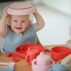 A smiling baby with a pink plate on its head sits at a table with baby feeding dishes.