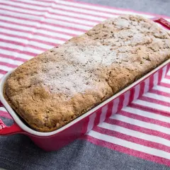 Pyszny chleb orkiszowo żytni na drożdżach, świeżo upieczony, w czerwonej formie Staub, na tle pasiastego obrusu.