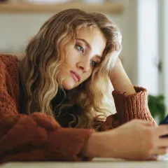 Młoda kobieta w brązowym swetrze, z ręką opartą o głowę, patrzy na telefon. Jej wyraz twarzy sugeruje anhedonię, brak radości z życia.