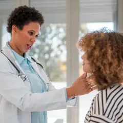 Lekarka bada szyję pacjentki, która ma charakterystyczną fryzurę. To ważny moment w diagnostyce, być może związany z problemami z tarczycą, gdzie kluczowe jest badanie palpacyjne, a czasem i leczenie z wykorzystaniem preparatów takich jak Theospirex.