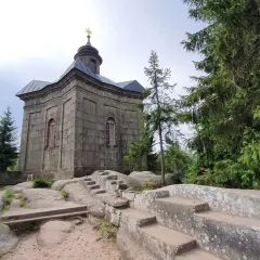 Kamienna kaplica na skale, otoczona drzewami. Broumowskie ściany budowli wznoszą się ku niebu, a kamienne schody prowadzą do jej wnętrza.