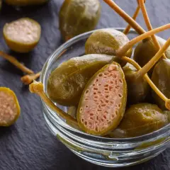 Zielone kapary w szklanej miseczce, jeden przekrojony ukazuje wnętrze. To właśnie kapary, co to za smak!