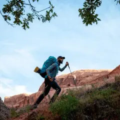 Hombre con mochila y bastones de senderismo sube una ladera rocosa bajo un cielo azul.