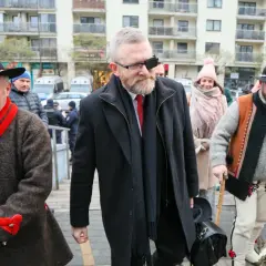 Grzegorz Braun w czarnym płaszczu i z opaską na oku, w otoczeniu mężczyzn w tradycyjnych strojach.