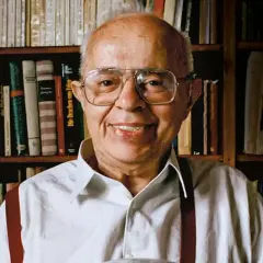 Uśmiechnięty Stanisław Lem, autor powieści Solaris, siedzi przed regałami pełnymi książek.