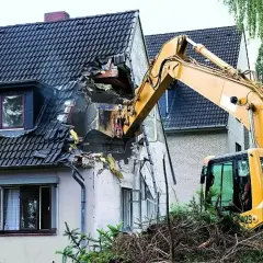 Żółta koparka prowadzi rozbiórkę dachu domu, niszcząc jego konstrukcję.