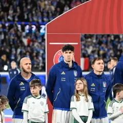 Piłkarze Włoch i Serbii stoją w rzędzie, dzieci w strojach kibicują.