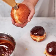 Eine Hand füllt einen Boston Cream Donut mit cremiger Füllung. Daneben liegen bereits glasierte Donuts.