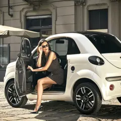 Elegancka kobieta w okularach przeciwsłonecznych wysiada z białego auta Microcar. To idealne auto bez prawa jazdy na miejskie wypady.