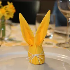 Gul servettvikning påsk, formad som en kanin, står på en tallrik. Bakom syns påskliljor och glas.