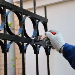 Ręka w rękawicy maluje czarną farbą metalową, ozdobną kratę. Farba, być może na bazie nafty, nadaje jej nowy, lśniący wygląd.