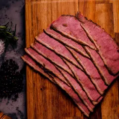 Cienkie plastry różowego pastrami na desce, obok przyprawy. Zastanawiasz się, pastrami co to? To aromatyczna, peklowana wołowina.