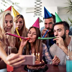 Grupa przyjaciół świętuje 40 urodziny w domu, dmuchając w trąbki i robiąc selfie z tortem.