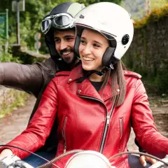 Pareja en moto, él con casco y gafas, ella con casco blanco y chaqueta roja, toman una selfie con el móvil. ¡Manos libres, casco moto legal y a disfrutar del viaje!