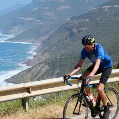 Man on a bike tour in Sardinia, cycling along a scenic coastal road with the blue sea and green hills.