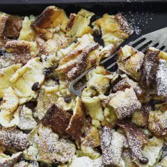 Kaiserschmarrn aus dem Backofen auf einem Blech, mit Puderzucker bestäubt und Rosinen. Ein Gabel-Schaber liegt bereit.