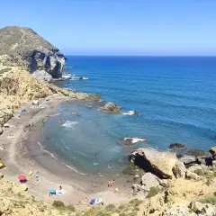 Vista aérea de **playas nudistas Cabo de Gata**. Arena oscura, aguas turquesas y formaciones rocosas. Gente disfrutando del sol y el mar.