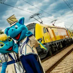 Dwa niebieskie smoki w strojach pozują przy żółtym pociągu. To pociąg do Energylandii, gotowy na podróż pełną wrażeń!