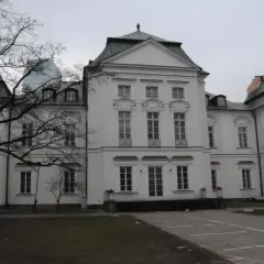 Pałac Przebendowskich, biała, elegancka budowla z zielonym dachem, otoczona drzewami i nowoczesnymi budynkami.