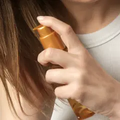 Delikatne olejowanie włosów rozjaśnianych złotym serum. Dłoń aplikuje produkt na brązowe pasma, przywracając im blask i nawilżenie.