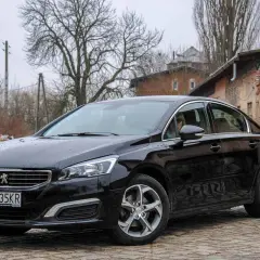 Czarny Peugeot 508 2.0 HDI na brukowanej drodze, w tle zniszczone budynki i drzewa.