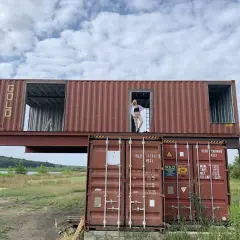Kobieta stoi w drzwiach domu z kontenera morskiego, który jest częścią nietypowej budowli.
