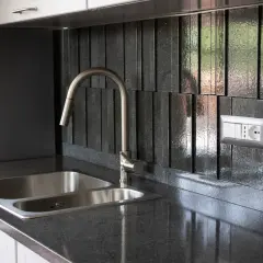 Top cucina in pietra lavica: pro e contro. Un lavello doppio in acciaio inox e un rubinetto moderno su un piano scuro e un rivestimento a listelli verticali.