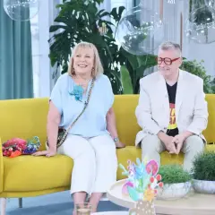 Uśmiechnięta para siedzi na żółtej kanapie. Pan Tik Tak w jasnej marynarce i kolorowym T-shircie.
