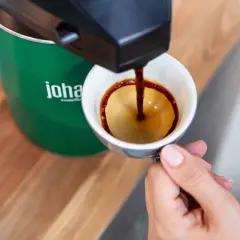 En hand håller en kopp medan kaffe rinner ner från en maskin. Frågan om hur många matskedar kaffe per kopp är relevant för att få den perfekta smaken.
