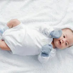 Malutki noworodek w białym body i niebieskich skarpetkach, z rękawiczkami na rączkach. Wygląda, jakby zastanawiał się, kiedy wreszcie zrobi kupę.