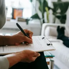 Osoba pisze na kartce na klipie, w tle kanapa i roślina.