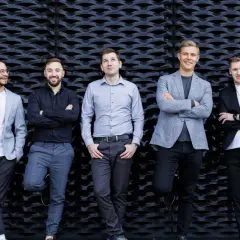 Pięciu mężczyzn w eleganckich strojach, idealnych do sesji fotografia biznesowa, pozuje na tle czarnej, geometrycznej ściany.