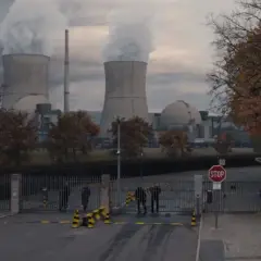 Mroczna elektrownia z chmurami pary nad chłodniami. Strażnicy przy bramie, nie wiadomo, kto jest kim.