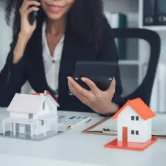 Agent immobilier au téléphone, calculatrice en main, devant des maquettes de maisons. Elle prépare une vente à terme.