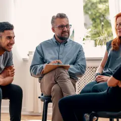 Grupa ludzi na sesji terapeutycznej. Mężczyzna z notatkami prowadzi rozmowę, a uczestnicy słuchają. Może uczą się języka francuskiego?