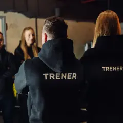Trzech trenerów w czarnych bluzach z napisami "TRENER" i "TRENERKA" stoi w siłowni.